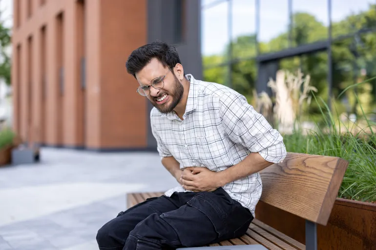 Contorted from severe pain, a young Indian man sits on a bench in the street and holds his stomach with his hands.; Shutterstock ID 2511048691; purchase_order: aj; job: ; client: ; other: